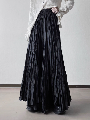 A person wearing a long black pleated maxi skirt with a gathered elastic waistband, styled with a white button-up shirt and black shoes, standing against a plain light gray wall.