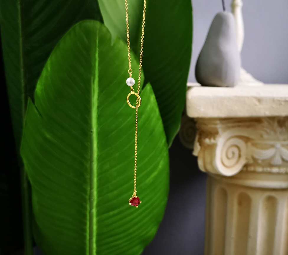 Gold necklace with a red pendant and white pearl on a green leaf background