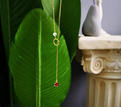 Gold necklace with a red pendant and white pearl on a green leaf background