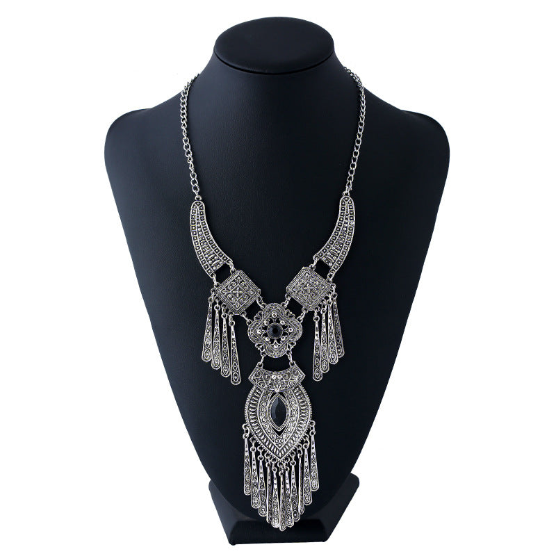 A silver-toned statement necklace with intricate filigree patterns and dangling fringe, displayed on a black jewelry bust against a white background.