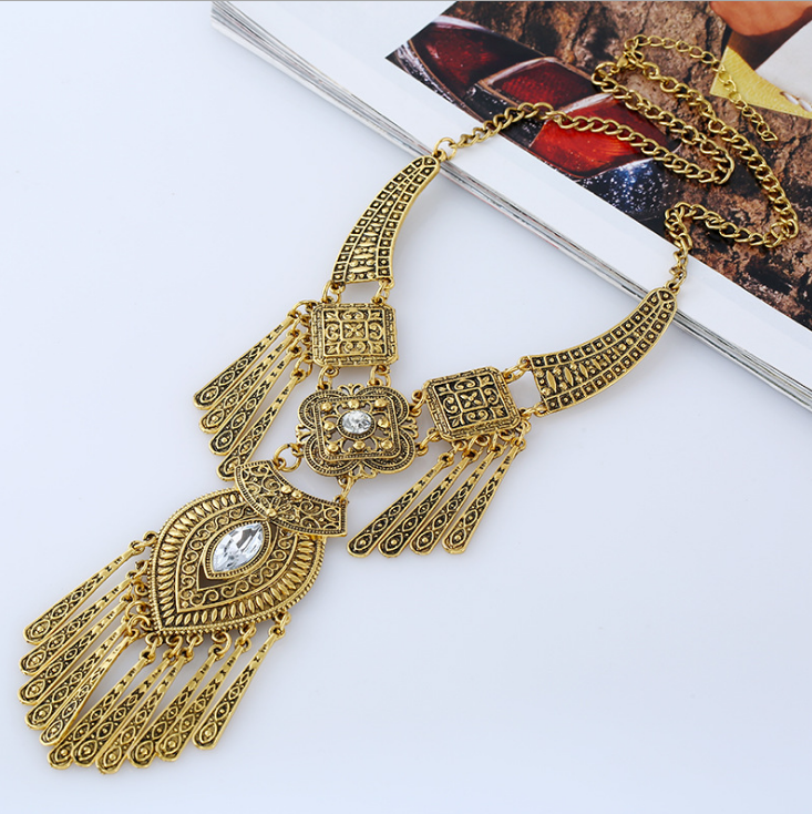 An ornate gold-toned statement necklace with intricate filigree detailing, a central clear rhinestone, and dangling fringe, displayed on a white surface with an open magazine.