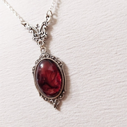 An antique-style silver pendant necklace featuring an oval deep red gemstone, displayed on a textured off-white fabric background.