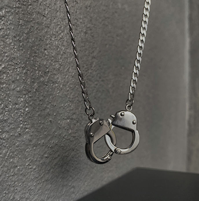 A silver-toned metal chain necklace with a pendant shaped like two interlocking handcuffs, photographed against a textured gray wall.