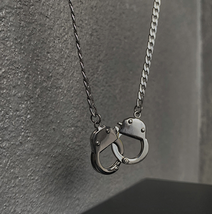 A silver-toned metal chain necklace with a pendant shaped like two interlocking handcuffs, photographed against a textured gray wall.