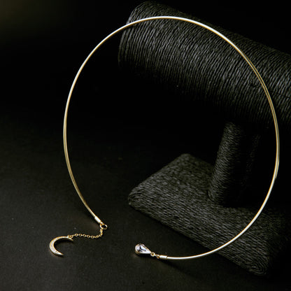 A gold-toned open circle choker necklace with a crescent moon charm and a teardrop crystal accent, displayed on a dark textured stand against a black background.