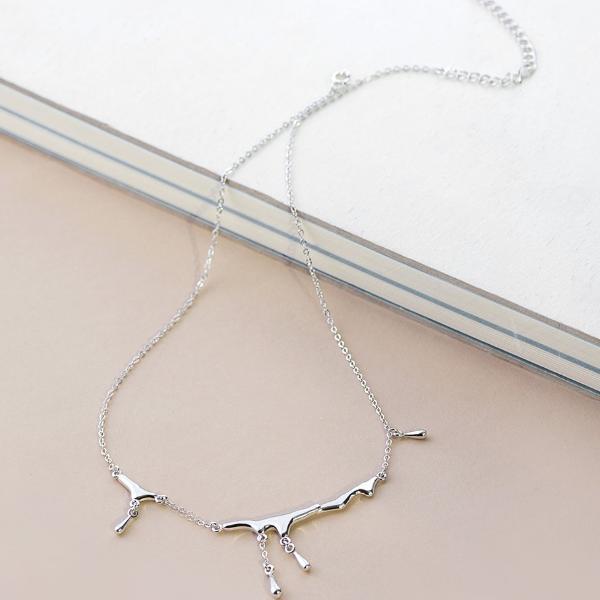 A delicate silver necklace with a minimalist abstract branch or tree-like pendant, resting on a light beige surface next to a white book.