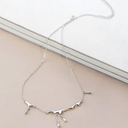 A delicate silver necklace with a minimalist abstract branch or tree-like pendant, resting on a light beige surface next to a white book.